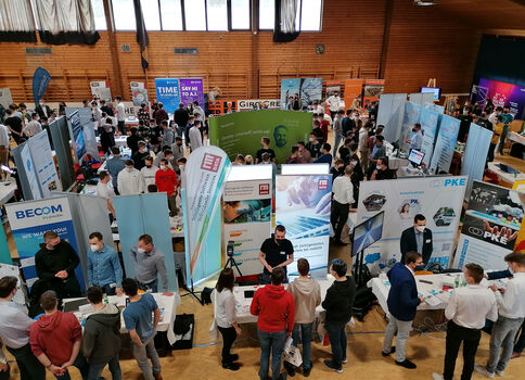 Eine große Turnhalle voller Messestände, Banner und Informationsstände; Besucher erkunden Stände, sprechen mit Ausstellern und sammeln Informationen.