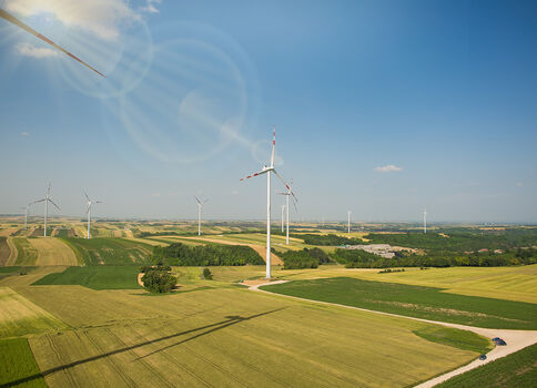 Weite Agrarlandschaft mit mehreren Windrädern auf Feldern; eine kurvige Straße führt durchs grüne Land, darüber ein blauer Himmel.