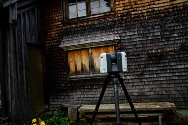 Ein Vermessungsgerät (Totalstation) auf einem Stativ steht vor einem alten Holzhäuschen.