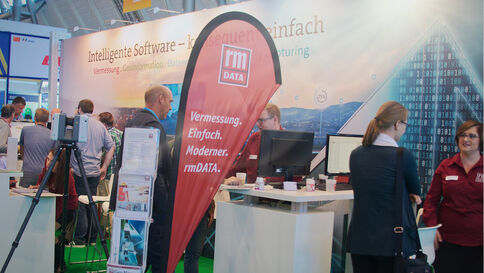 Ausstellungsstand von rmDATA auf einer Messe. Besucher arbeiten an Computern, hinter dem Stand hängt ein Banner mit dem rmDATA-Logo und dem Slogan "Vermessung. Einfach. Modern."