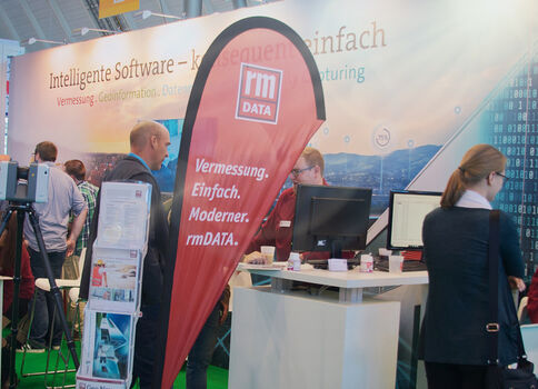Ausstellungsstand von rmDATA auf einer Messe. Besucher arbeiten an Computern, hinter dem Stand hängt ein Banner mit dem rmDATA-Logo und dem Slogan "Vermessung. Einfach. Modern."