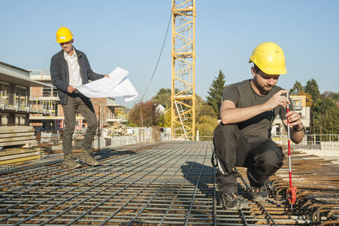 Zwei Bauarbeiter mit gelben Helmen arbeiten auf einer Baustelle. Einer hält Baupläne, der andere misst mit einem Messwerkzeug über einem Stahlbewehrungsnetz; im Hintergrund steht ein Baukran.
