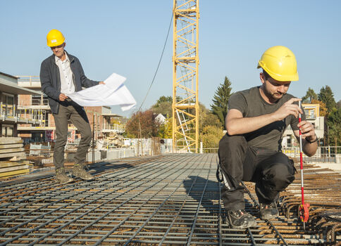 Zwei Bauarbeiter mit gelben Helmen arbeiten auf einer Baustelle. Einer hält Baupläne, der andere misst mit einem Messwerkzeug über einem Stahlbewehrungsnetz; im Hintergrund steht ein Baukran.
