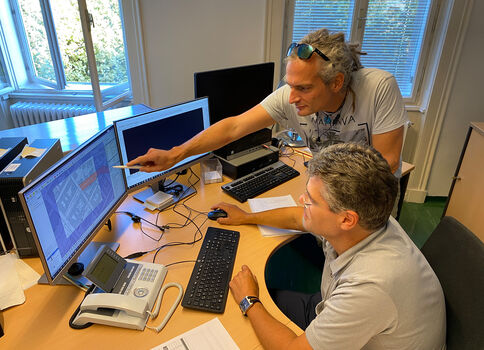 Zwei Männer arbeiten in einem Büro an einem Schreibtisch mit mehreren Monitoren. Einer steht und zeigt mit einem Stift auf den Bildschirm, der andere sitzt und schaut aufmerksam zu. Im Hintergrund Fenster mit Vorhängen und weitere Bürotechnik.