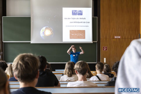 Vorlesung: Ein Dozent in blauem T‑Shirt steht vor einer Folienpräsentation in einem Hörsaal; auf der Folie sind ein Erdbild, zwei Logos und Textzeilen zu sehen. Im Vordergrund sitzen Studierende und schauen zur Bühne.