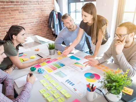 Fünf Personen sitzen um einen hellen Konferenztisch und arbeiten gemeinsam an einem Projekt. Auf dem Tisch liegen Post-its, Diagramme, Farbkarten, ein Laptop und Marker; die Gruppe diskutiert Inhalte und analysiert Diagramme.