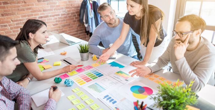 Groupe de cinq professionnels en réunion autour d'une table, analysant des graphiques et des post-it colorés. Ils discutent et prennent des notes, avec un ordinateur portable et des documents visibles, dans une salle lumineuse au mur en briques.