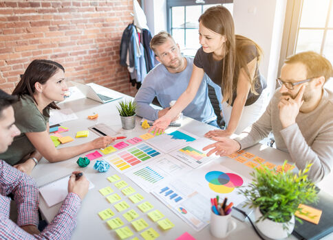 Fünf Personen sitzen um einen hellen Konferenztisch und arbeiten gemeinsam an einem Projekt. Auf dem Tisch liegen Post-its, Diagramme, Farbkarten, ein Laptop und Marker; die Gruppe diskutiert Inhalte und analysiert Diagramme.
