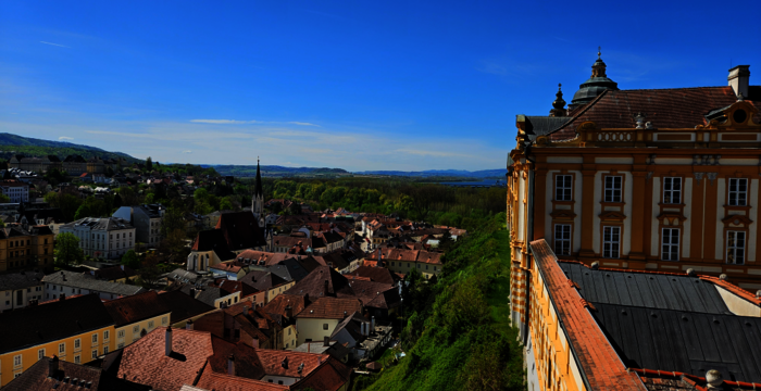 Ausblick über eine malerische Stadt mit roten Ziegeldächern, Kirchtürmen und engen Gassen. Rechts im Vordergrund ein prachtvolles Barockgebäude, links grüne Hügel und ein weiter blauer Himmel.