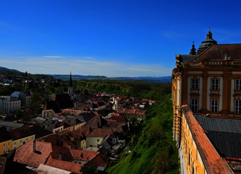 Ausblick über eine malerische Stadt mit roten Ziegeldächern, Kirchtürmen und engen Gassen. Rechts im Vordergrund ein prachtvolles Barockgebäude, links grüne Hügel und ein weiter blauer Himmel.