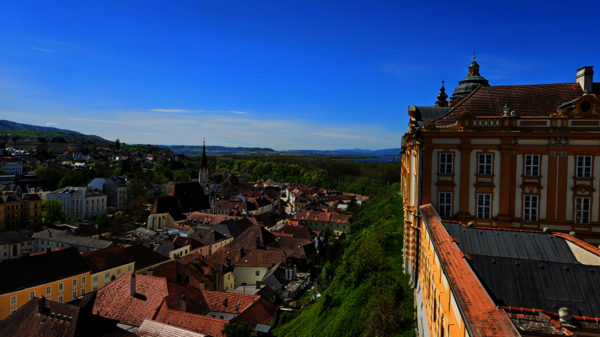 Ausblick über eine malerische Stadt mit roten Ziegeldächern, Kirchtürmen und engen Gassen. Rechts im Vordergrund ein prachtvolles Barockgebäude, links grüne Hügel und ein weiter blauer Himmel.