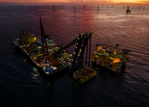 Offshore-Bohrplattform im Meer bei Sonnenuntergang. Gelbe Strukturteile und ein großer Kran ragen aus dem Wasser, Beleuchtung leuchtet. Im Hintergrund sind weitere Ölplattformen und Pfähle zu sehen. Ruhiges Wasser spiegelt den warmen, orangefarbenen Himmel.