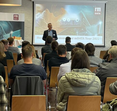 Ein Redner präsentiert vor einer Zuhörerschaft in einem Konferenzraum. Eine Projektion zeigt die Willkommensfolie der mDATA Tour 2022 mit dem rmDATA-Logo; das Publikum sitzt in Stuhlreihen und schaut zur Bühne.