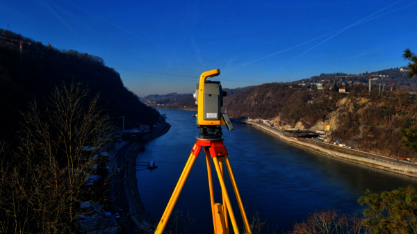 A yellow surveying instrument on an orange tripod stands on a riverside overlook, surveying a winding river between forested hills with a distant hillside town under a clear blue sky.