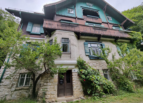 Grande maison de style chalet, façade en pierre au rez-de-chaussée avec boiseries sur les étages supérieurs, volets verts et balcon en bois, entourée d'arbres et d'arbustes.