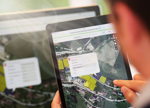 Auf einem Tablet ist eine Kartenansicht mit Satellitenbild zu sehen; über der Karte liegt ein Informationsfenster.