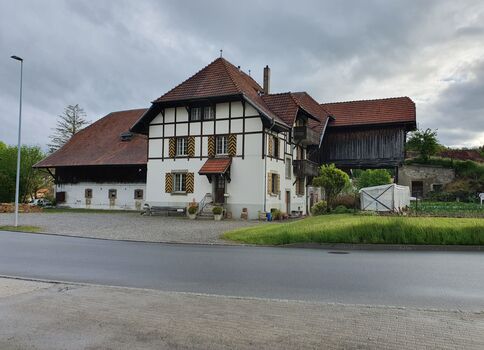 Ferme traditionnelle à colombages près d'une route: grande maison blanche à ossature en bois avec volets et toit rouge, flanquée d’un long bâtiment blanc et d’une grange en bois surélevée. Ciel nuageux, pelouse et gravier devant.