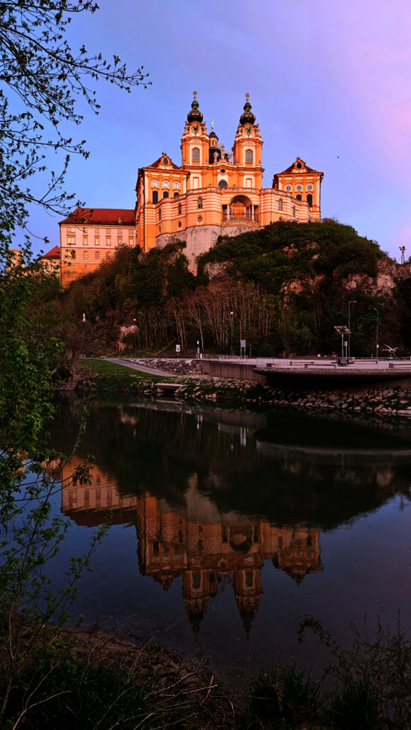 Eine prunkvolle Barockkirche mit zwei Türmen steht auf einem Hügel über einem Fluss. Der Bau und umliegende Bäume spiegeln sich im ruhigen Wasser, während der Abendhimmel rosa-violett leuchtet.