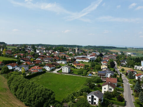 Luftaufnahme eines kleinen Dorfes mit roten Dächern, Einfamilienhäusern und Gärten, verteilten Straßen und einer Kirchturmsilhouette in der Mitte. Rundherum Felder, Wiesen und Bäume prägen die ländliche Landschaft.
