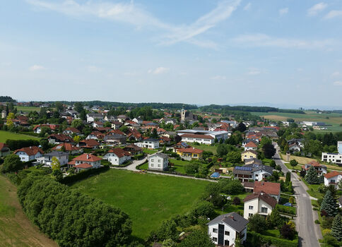 Luftaufnahme eines kleinen Dorfes mit roten Dächern, Einfamilienhäusern und Gärten, verteilten Straßen und einer Kirchturmsilhouette in der Mitte. Rundherum Felder, Wiesen und Bäume prägen die ländliche Landschaft.