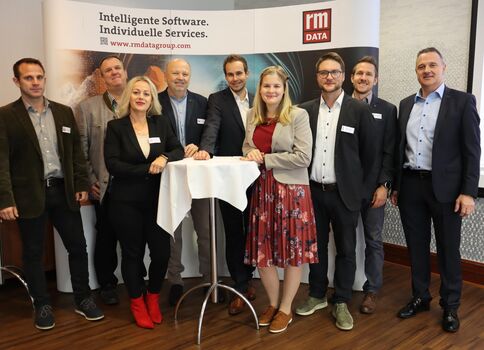 Eine Gruppe beruflich gekleideter Menschen posiert an einem Stehtisch bei einer Firmenveranstaltung. Im Hintergrund ist ein Banner mit dem Text 'Intelligente Software. Individuelle Services.' und dem rmData-Logo zu sehen; es wirkt wie ein Networking- oder Präsentationsmoment.