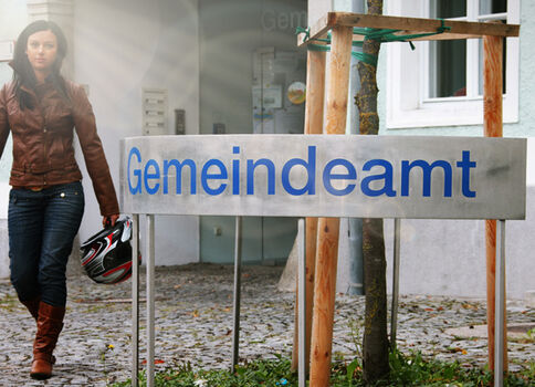 Eine Frau in brauner Jacke geht am Gemeindeamt vorbei; sie trägt Jeans, Stiefel und hält einen Motorradhelm in der Hand.