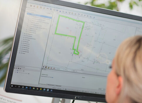 Ein Computerbildschirm zeigt GIS‑Karten-Software mit einer grün markierten Route auf einer Karte; im Vordergrund sitzt eine Person mit blondem Haar und schaut auf den Bildschirm.