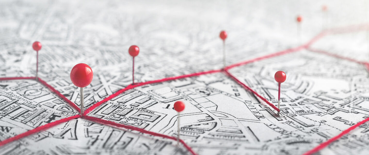 Nahaufnahme einer Karte mit grauen Straßenlinien. Mehrere rote Stecknadeln markieren Standorte, die durch rote Linien zu einem Netzwerk verbunden sind – eine grafische Darstellung von Verbindungen oder Routen.