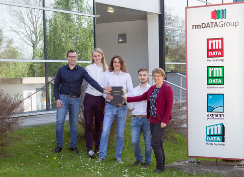 Fünf Personen stehen vor einem Bürogebäude und posieren mit einem aufgeschlagenen Tablet. Rechts daneben befindet sich ein rmDATA Group Schild mit Logos der Tochterunternehmen.