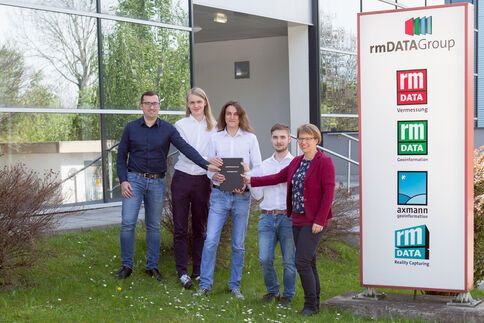 Fünf Personen stehen vor einem Bürogebäude und posieren mit einem aufgeschlagenen Tablet. Rechts daneben befindet sich ein rmDATA Group Schild mit Logos der Tochterunternehmen.