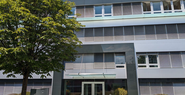 Mehrstöckiges Bürogebäude aus Glas und Beton mit auffälliger Eingangstür unter einem Vordach. Links steht ein großer Baum, rechts niedrige Sträucher. Die Fassade zeigt horizontale Fensterläden und graue Fassadenelemente; der Himmel ist blau.