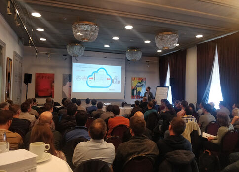 Une salle de conférence bondée où un présentateur se tient près d’un écran projetant une diapositive montrant un nuage bleu; le public est assis et écoute la présentation.