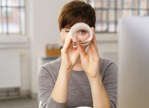 Eine Person mit kurzen Haaren schaut durch einen weißen Ring, den sie mit den Händen formt, und lächelt. Im Vordergrund ein Computer; im Hintergrund ein helles Büro mit Fenstern.