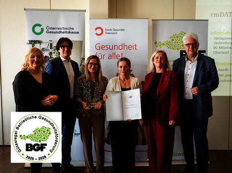 Sechs Personen in Business-Outfits posieren vor Bannern der österreichischen Gesundheitsorganisationen. Eine Frau in der Mitte hält eine geöffnete Urkunde; Logos wie ÖGK und Fonds Gesundheit sichtbar. Unten links ein runder BGF-Aufkleber. Die Szene wirkt wie eine formelle Zertifikatsübergabe.
