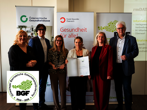 Sechs Personen in Business-Outfits posieren vor Bannern der österreichischen Gesundheitsorganisationen. Eine Frau in der Mitte hält eine geöffnete Urkunde; Logos wie ÖGK und Fonds Gesundheit sichtbar. Unten links ein runder BGF-Aufkleber. Die Szene wirkt wie eine formelle Zertifikatsübergabe.