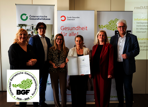 Sechs Personen in Business-Outfits posieren vor Bannern der österreichischen Gesundheitsorganisationen. Eine Frau in der Mitte hält eine geöffnete Urkunde; Logos wie ÖGK und Fonds Gesundheit sichtbar. Unten links ein runder BGF-Aufkleber. Die Szene wirkt wie eine formelle Zertifikatsübergabe.