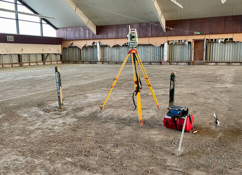Intérieur d'une grande salle vide. Au centre, un trépied jaune porte un instrument de relevé; à côté se trouvent un sac rouge et une petite boîte bleue. Deux piquets verts dans le sol; en arrière-plan, mur avec panneaux métalliques et grandes fenêtres.