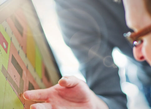 Eine Person mit Brille schaut auf einen Computerbildschirm, der ein farbiges Layout mit quadratischen Blöcken in Pink, Grün und Gelb zeigt; eine Hand zeigt auf das Display.