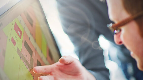 Produkteinführungsschulung Eine Person mit Brille schaut auf einen Computerbildschirm, der ein farbiges Layout mit quadratischen Blöcken in Pink, Grün und Gelb zeigt; eine Hand zeigt auf das Display.