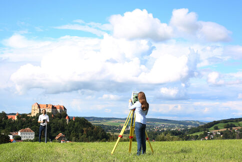 Zwei Personen arbeiten auf einer grünen Wiese: Eine bedient ein Vermessungsgerät auf einem Stativ, die andere steht daneben. Im Hintergrund erstreckt sich eine ländliche Landschaft mit einer Burg und Dörfern unter einem blauen Wolkenhimmel.