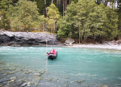 Eine Person in roter Jacke paddelt in einem kleinen aufblasbaren Boot auf einem türkisfarbenen Fluss, umgeben von felsigen Ufern und Wald.