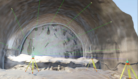 Innenansicht eines Tunnels im Rohbau. Vorne stehen drei Vermessungsstative mit gelb-roten Füßen, aus denen grüne Laserstrahlen durch den Tunnel ziehen und die Felswand ausleuchten.