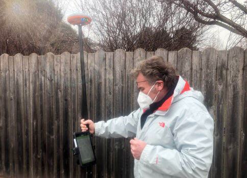 Ein Mann mit Mundschutz und hellgrauer Jacke steht draußen vor einem Holzzaun und bedient ein mobiles Messgerät an einem Stab mit Display.