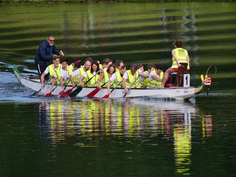 Drachenboot mit vielen Paddlern in gelben Rettungswesten, die synchron über einen ruhigen Fluss paddeln; am Heck steuert eine Person, am Bug sitzt ein Drachenkopf. Wasser spiegelt das Boot.