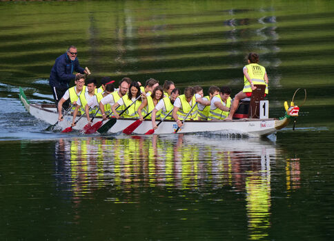 Drachenboot mit vielen Paddlern in gelben Rettungswesten, die synchron über einen ruhigen Fluss paddeln; am Heck steuert eine Person, am Bug sitzt ein Drachenkopf. Wasser spiegelt das Boot.