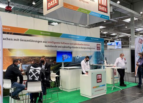 Auf einem Messestand präsentiert ein Aussteller Gesamtlösungen aus intelligenter Software und individuellen Services. Mehrere Besucher sitzen und stehen im Gespräch, hinter ihnen Informationsgrafiken. Über dem Stand hängen DATA-Schilder; die Szene wirkt wie eine Messehalle mit Monitoren und Präsentationsmaterial.