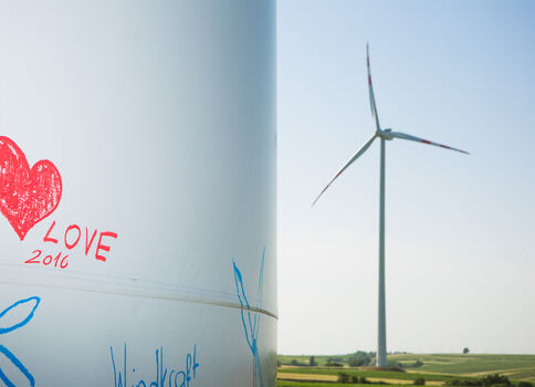 Eine Windkraftanlage steht in einer offenen Felderlandschaft; im Vordergrund eine siloförmige Struktur mit rotem Herz-Graffiti und der Aufschrift 'LOVE 2016', daneben blaue Blütenzeichnungen.