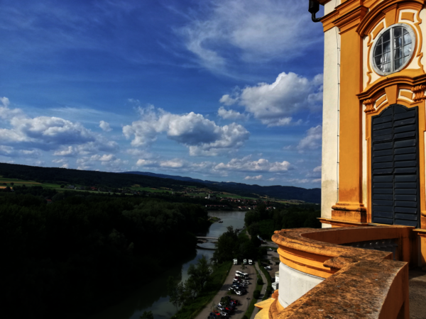 Ausblick von einer Terrassenkante eines gelb-orangefarbenen Barockgebäudes: ein Fluss schlängelt sich durch grüne Wiesen und Wälder, unter einem blauen Himmel mit Wolken.