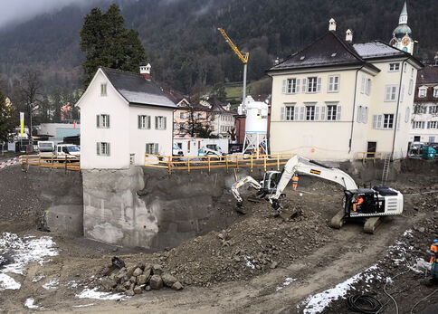 Eine Baustelle in einer kleinen Stadt: Ein Bagger gräbt an einer freigelegten Uferböschung, umgeben von weißen Häusern. Im Hintergrund ragt ein Baukran auf, Schutt und Schnee liegen auf dem Gelände, Absperrungen sind sichtbar.