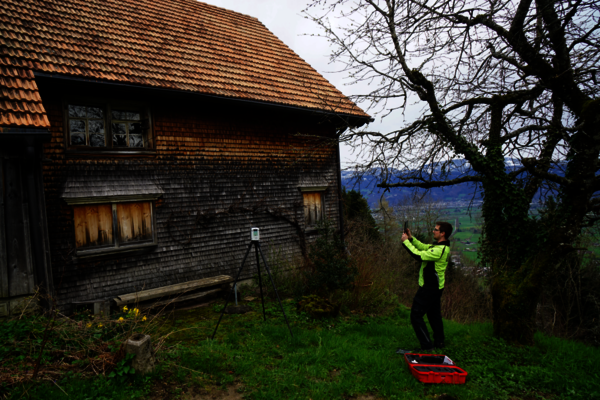 Eine Person in einer leuchtend gelben Jacke positioniert eine Kamera auf einem Stativ neben einem alten, holzverkleideten Haus mit rotem Ziegeldach; ein roter Werkzeugkasten liegt auf dem Gras, im Hintergrund eine ländliche Landschaft mit kahlen Bäumen.